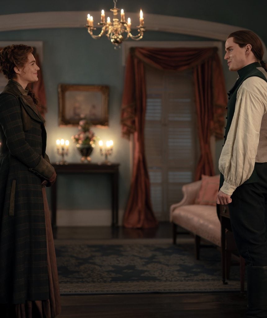 A woman and a man in period clothing stand facing each other in a dimly lit, elegantly furnished room with a chandelier and draped windows.