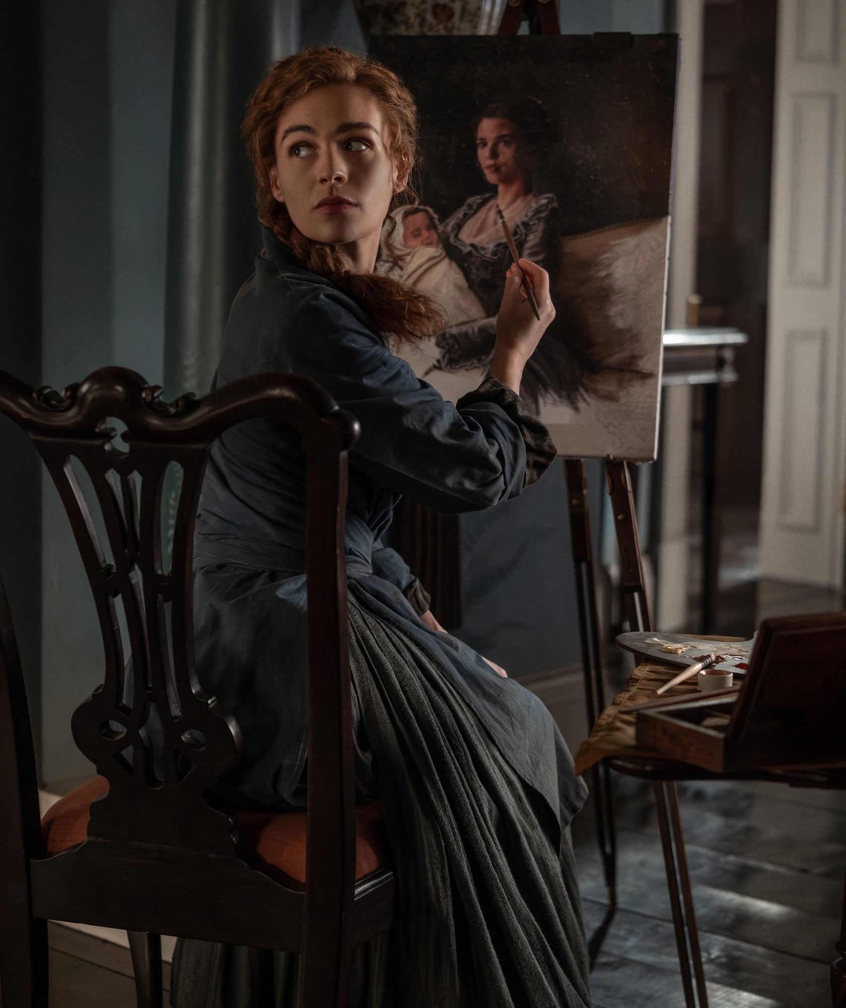 A woman in historical clothing sits on a chair, painting a portrait of a seated woman with a child on a canvas in an art studio.