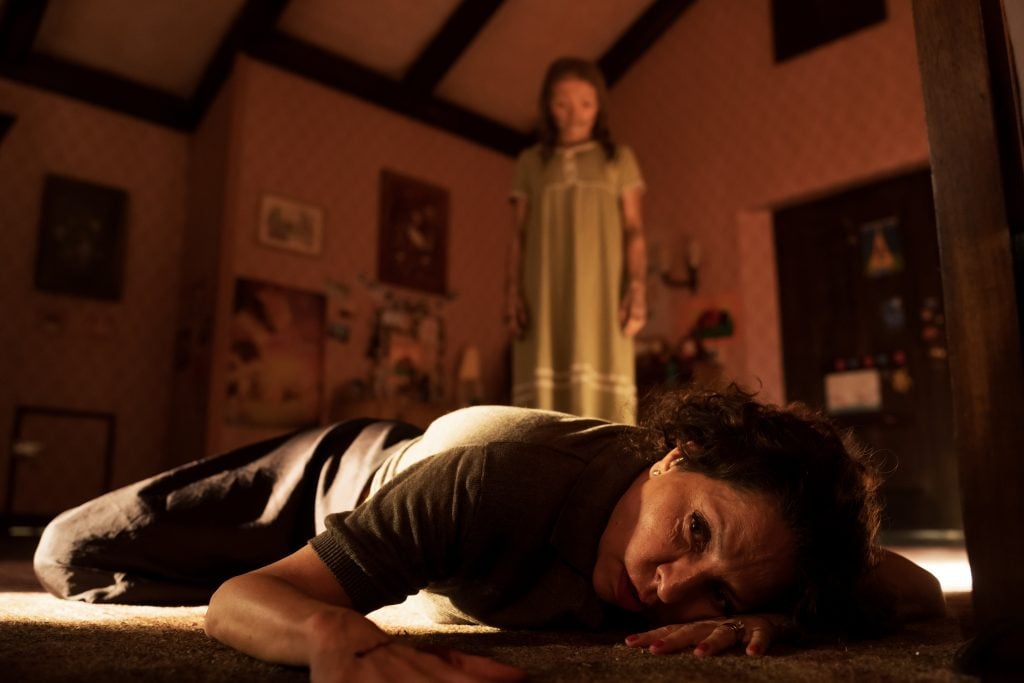 A woman lies on the floor looking distressed while a girl in a nightgown stands in the background of a dimly lit room.