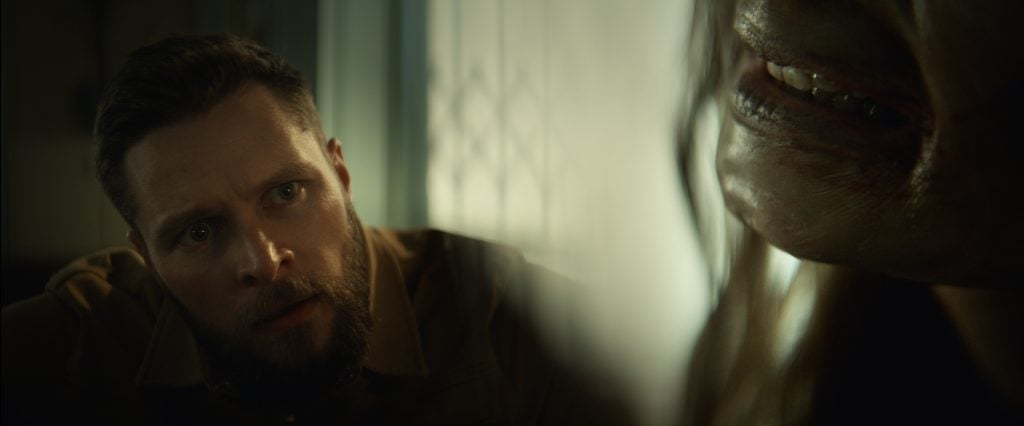 A man with a concerned expression looks toward a shadowy figure with decayed teeth and rough skin in a dimly lit room.