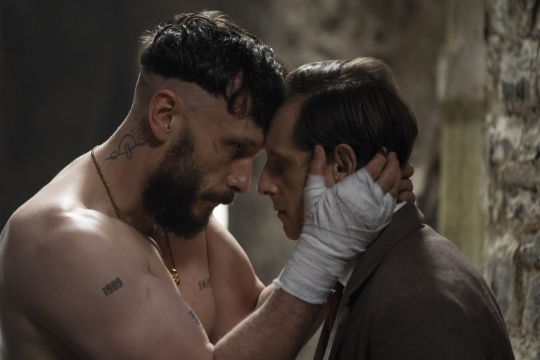 A shirtless man with tattoos holds a man's face close, both with serious expressions, in a dim, stone-walled room. The suited man's hand is wrapped in bandages.