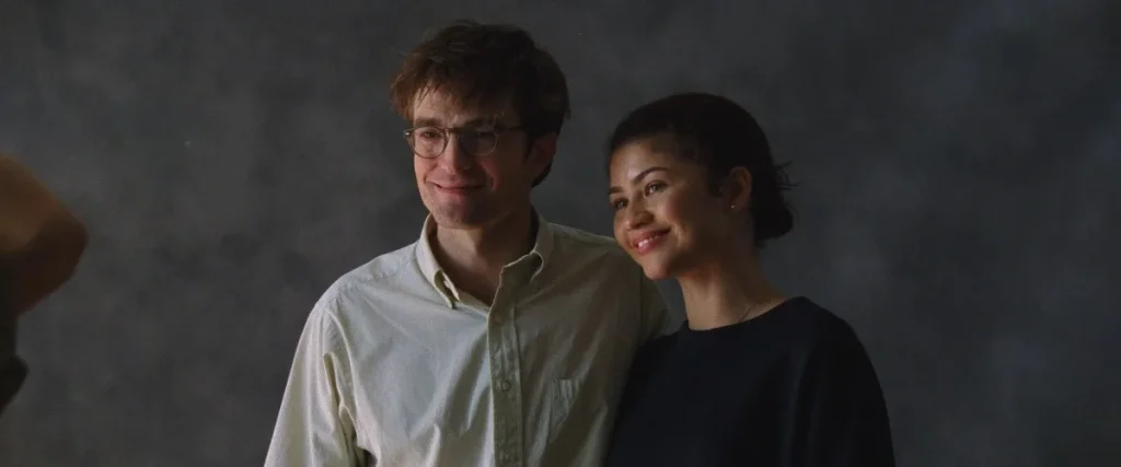 Two people stand side by side, smiling for a photo against a plain dark background. The person on the left wears glasses and a light shirt; the person on the right wears a dark top.