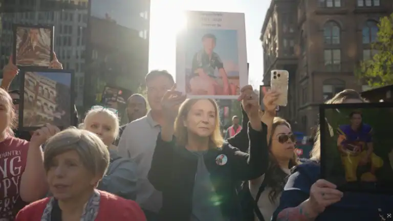 A group of people stand outdoors, holding framed photos and a sign with a young person's portrait and text, during a daylight gathering in an urban area.