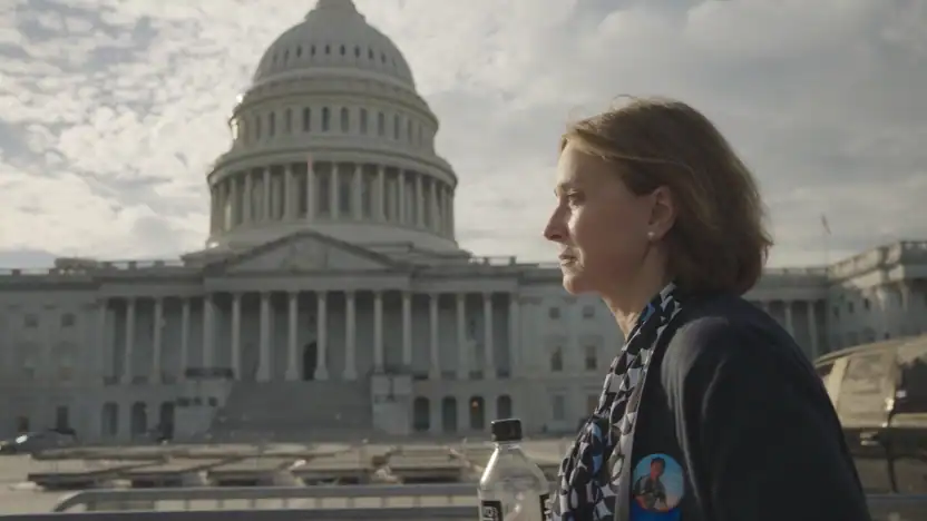 A woman stands outside the U.S. Capitol building, holding a bottle and wearing a scarf and a button with a person's face on it.