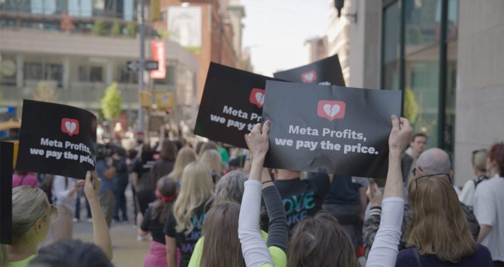 People at a protest hold signs that read "Meta Profits, we pay the price" with a broken heart icon, standing in an urban street.