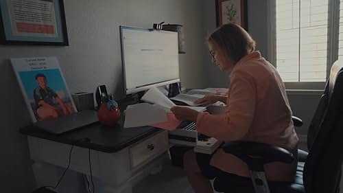A person sits at a desk working with papers and a laptop in a home office. A large monitor, framed photo, and window are visible in the background.