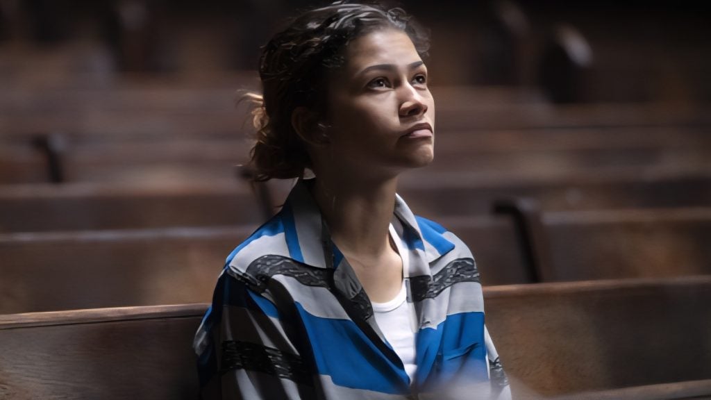 A young woman in a blue and gray striped shirt sits alone on a wooden bench in a dimly lit room, looking slightly upward with a serious expression.