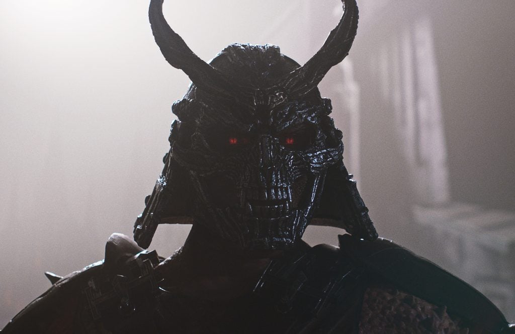 A figure wearing a dark, horned samurai helmet with glowing red eyes stands in a misty, dimly lit environment.