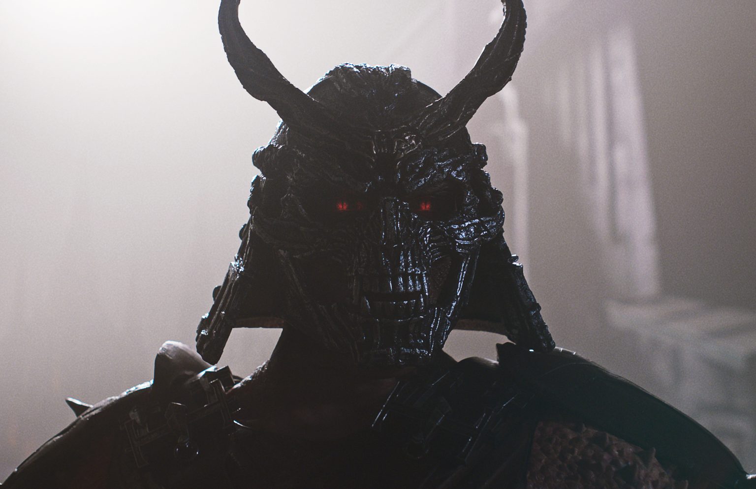 A figure wearing a dark, horned samurai helmet with glowing red eyes stands in a misty, dimly lit environment. Mortal Kombat II