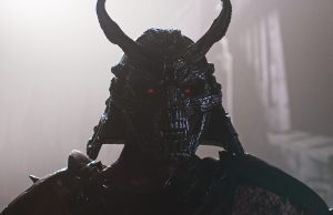 A figure wearing a dark, horned samurai helmet with glowing red eyes stands in a misty, dimly lit environment. Mortal Kombat II