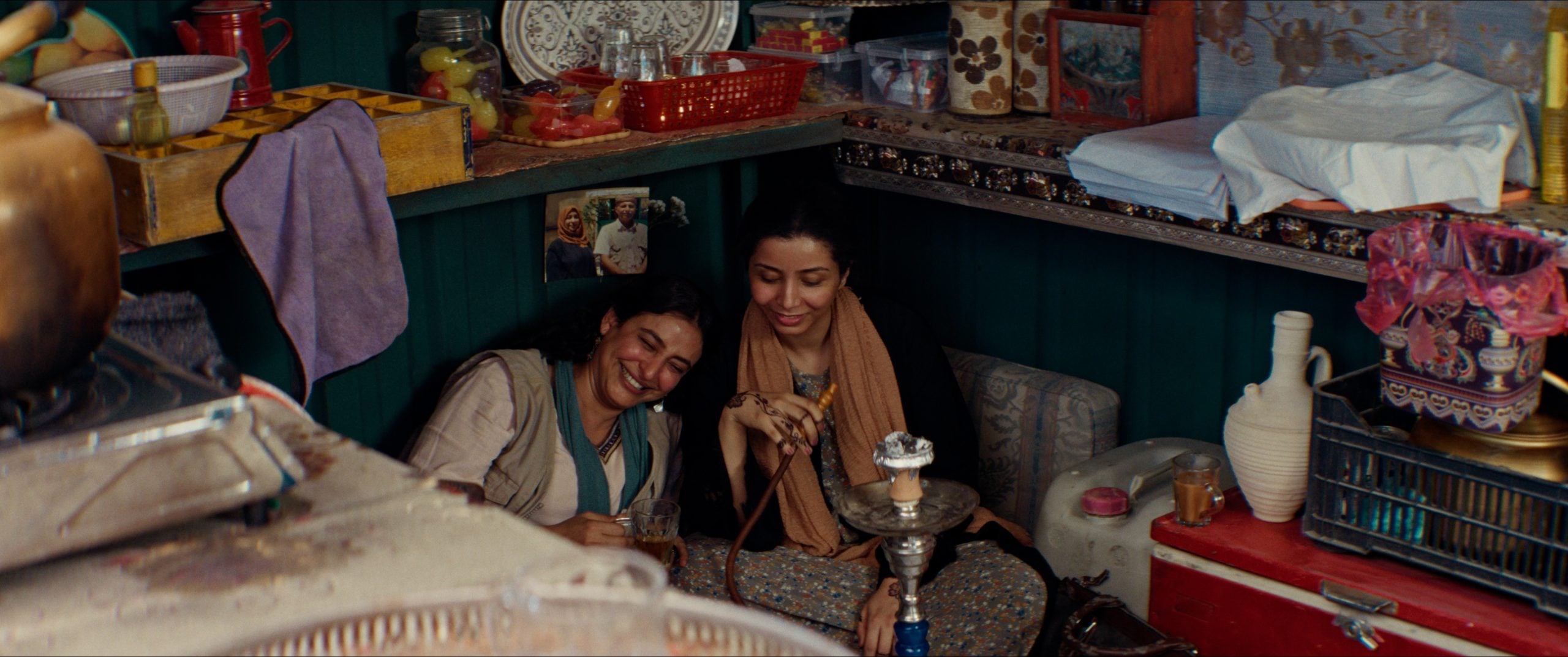 Two women sit on the floor of a cluttered room, smiling and sharing a hookah, with beverages and various items surrounding them.