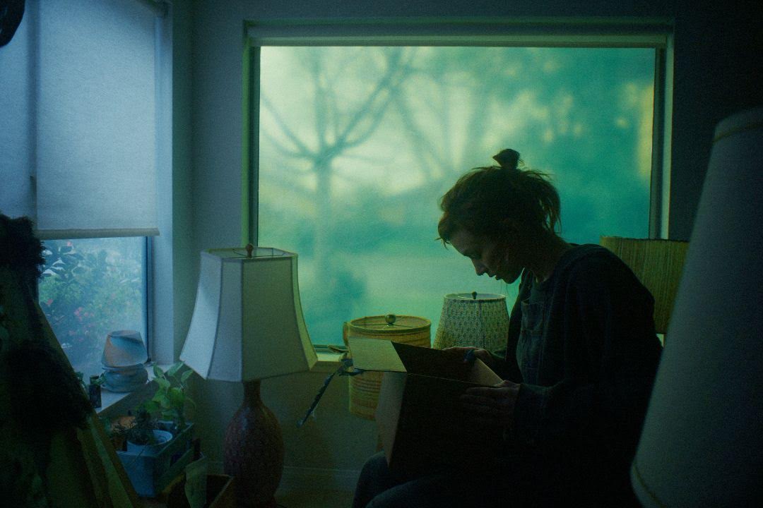 A person sits by a window in a dimly lit room, looking down into an open box. Outside, tree branches are visible through the greenish glass.