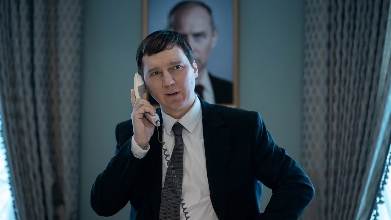 A man in a suit speaks on a landline phone, standing in an office with a portrait of another man hanging on the wall behind him.