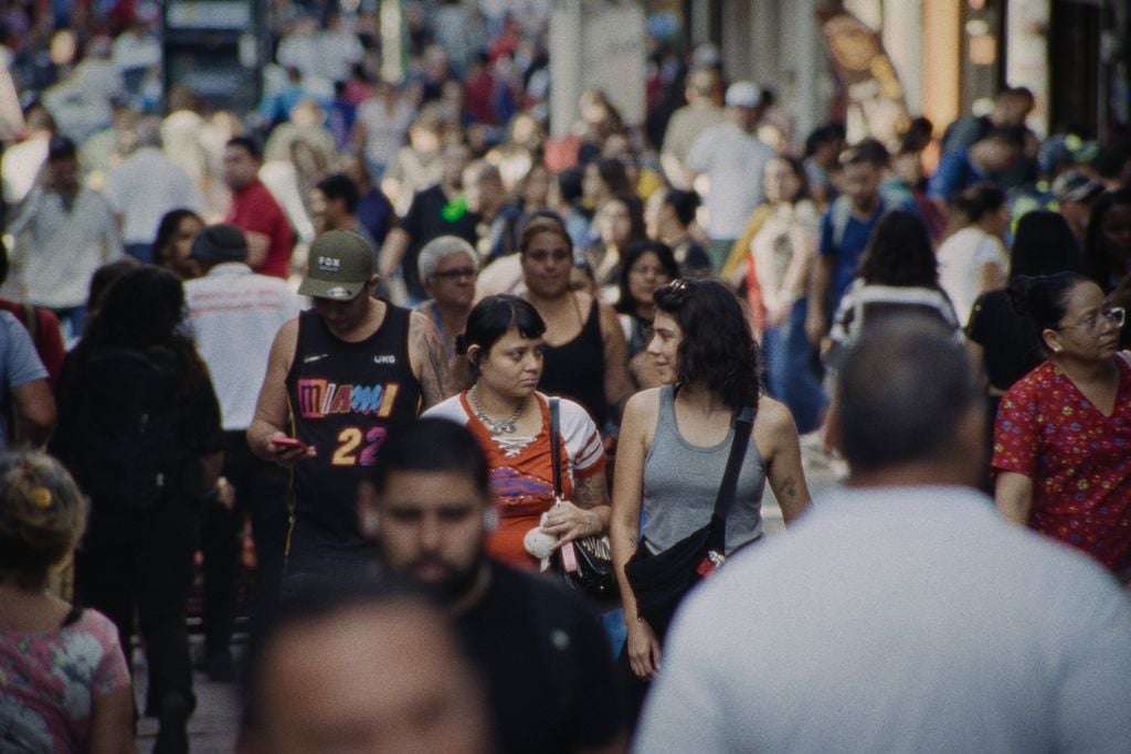 A busy city street filled with people walking in various directions, some talking, some looking at phones, and others passing by.