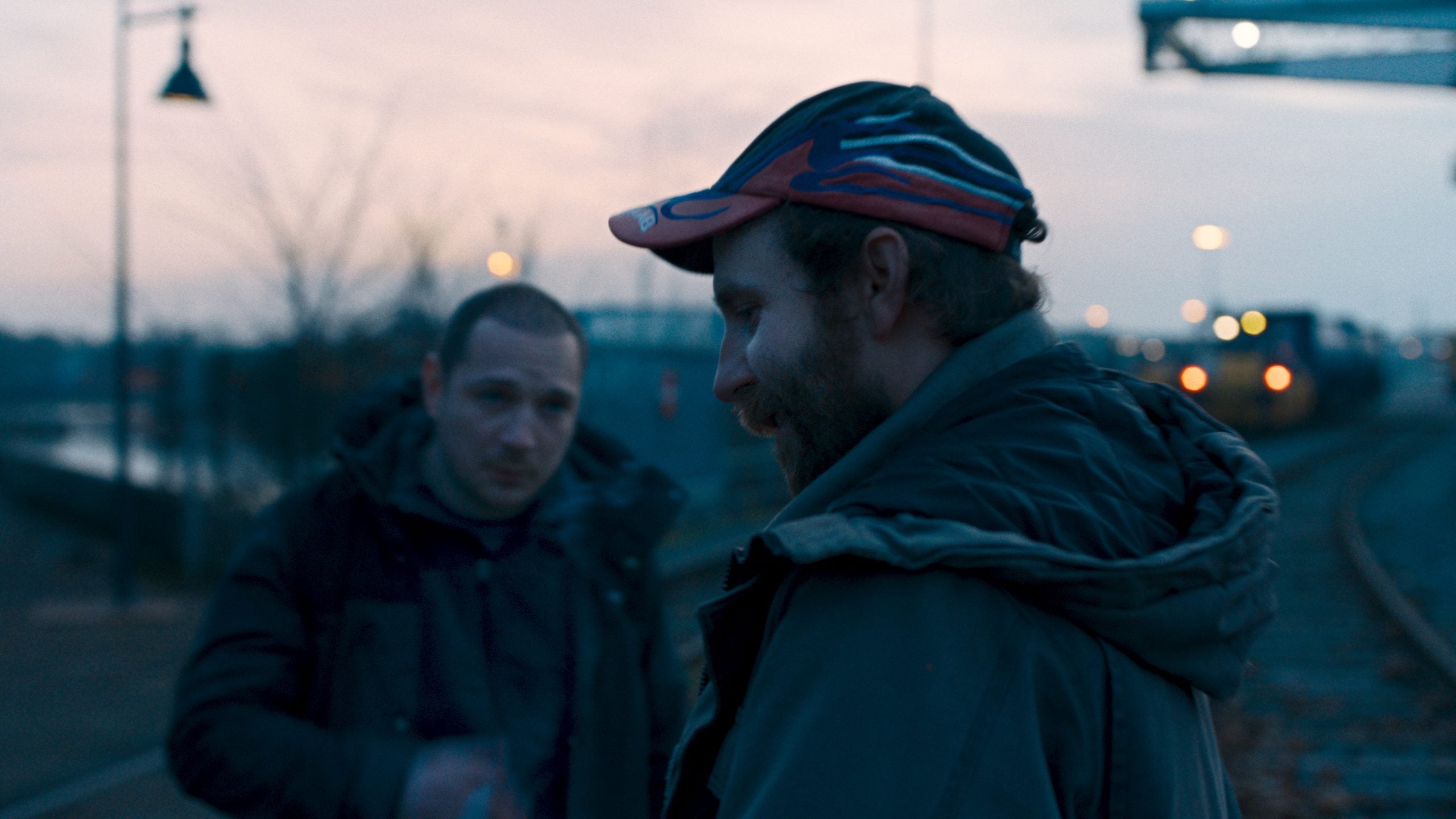 Two men stand outdoors near train tracks at dusk, one in the foreground wearing a cap and jacket, the other blurred in the background.