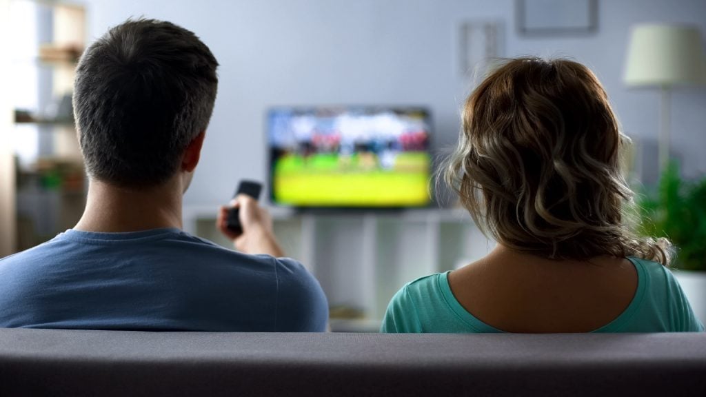 Two people sitting on a couch watching a sports game on a television; one person is holding a remote control.
