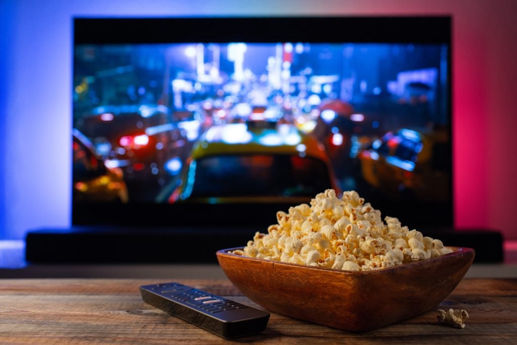 A bowl of popcorn and a TV remote on a wooden table in front of a television displaying a blurred city scene.