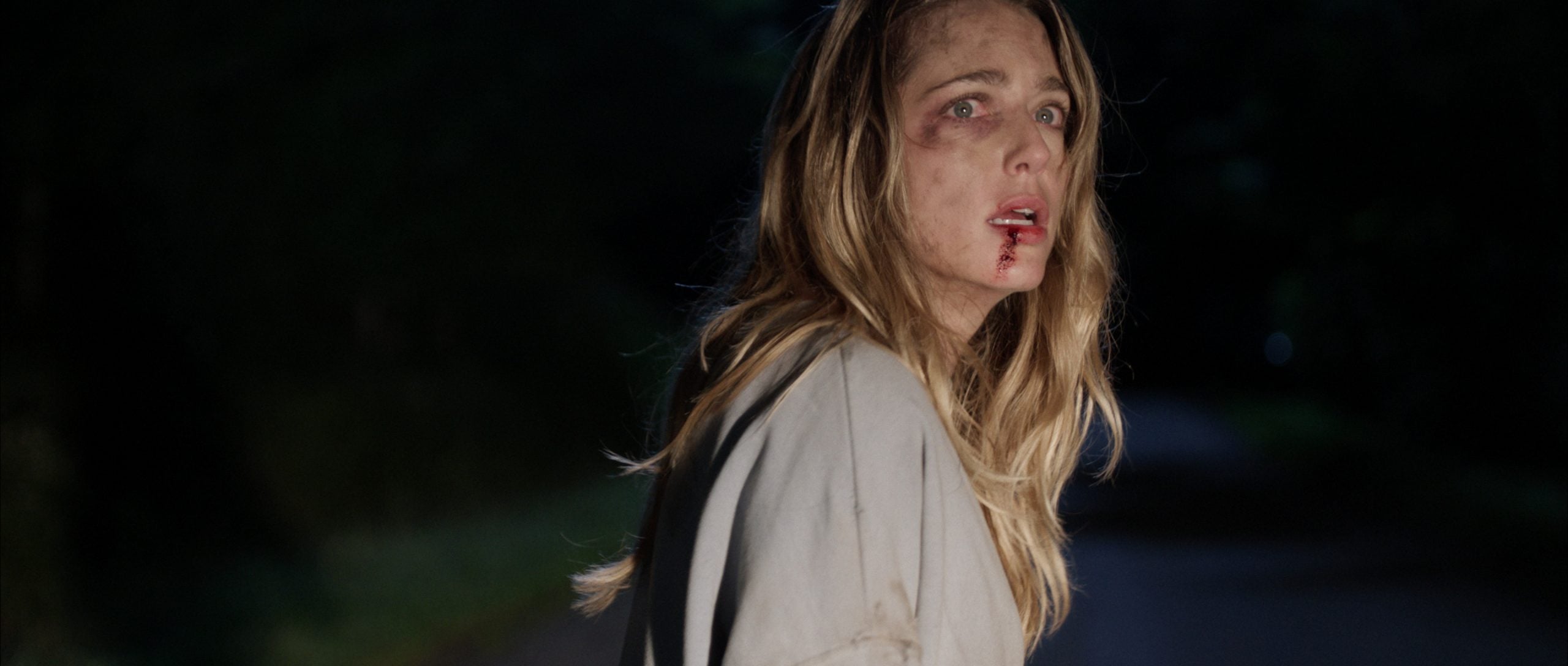 A woman with bruises and blood on her face stands outside at night, looking distressed and wearing a light-colored coat.