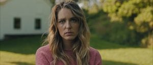 ‘Affection’ Blu-Ray Review – Jessica Rothe Elevates Uneven Memory Loss Thriller A woman with long blonde hair wearing a pink sweater stands outside on grass, looking directly at the camera with a neutral expression. A white building is in the background.