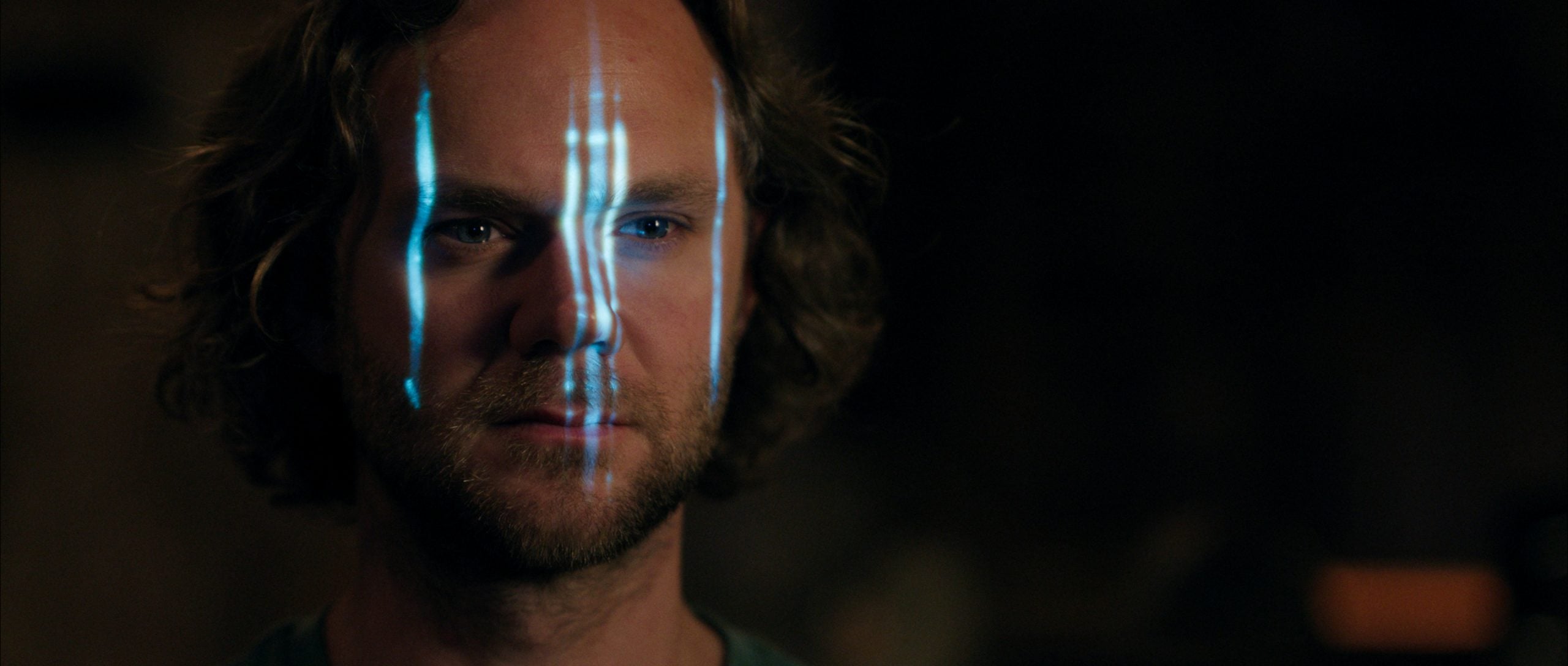 A man with shoulder-length hair has blue vertical light lines projected onto his face in a dark setting.