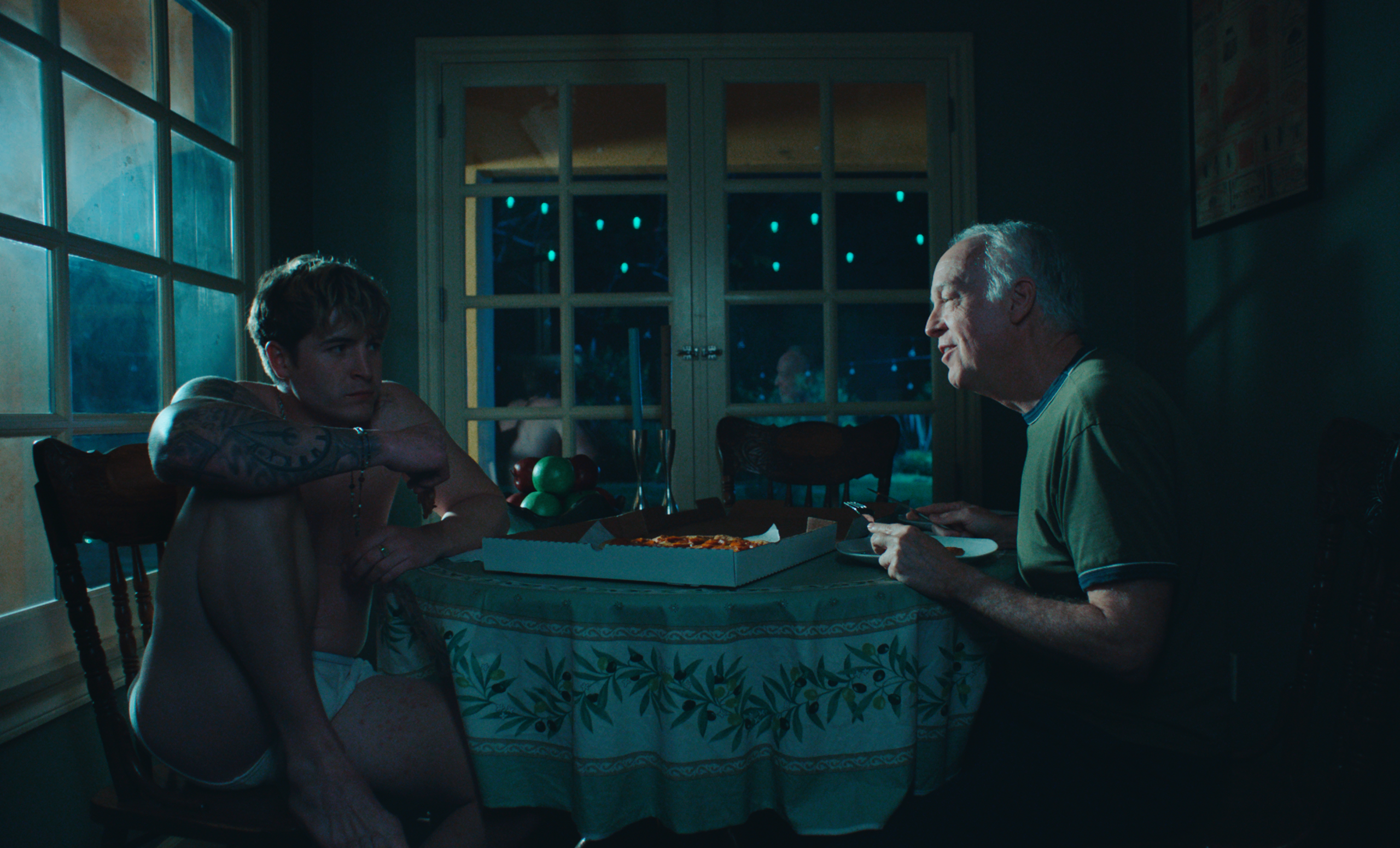 A young man in underwear sits on a chair with his knees up, while an older man eats pizza at a table in a dimly lit room.