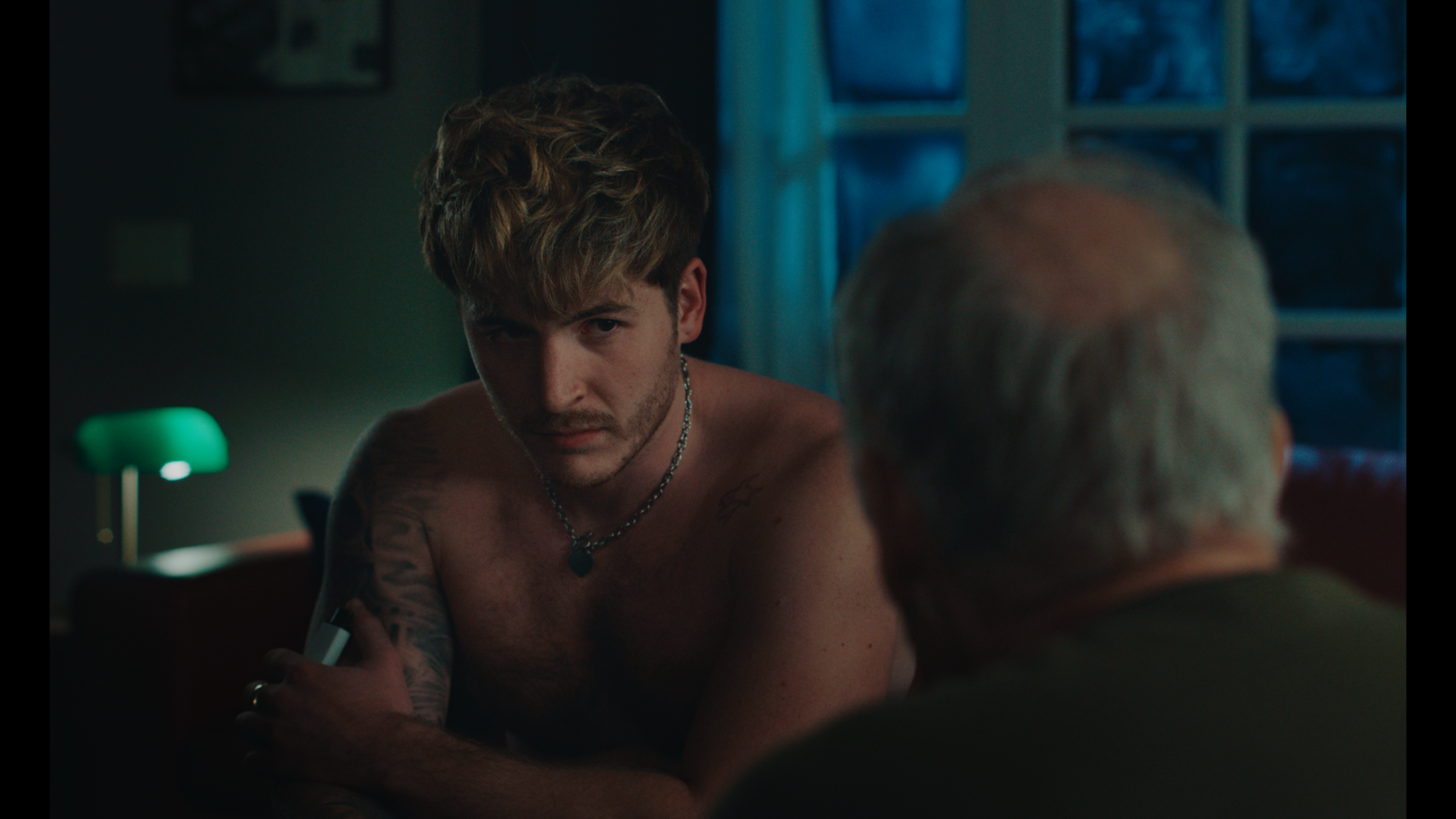 A shirtless young man with blonde hair and tattoos sits indoors at night, looking intently at an older man whose back is to the camera.