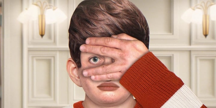 A person with a serious expression holds a hand over their face, with an eye drawn on the hand, against a background of white paneled walls and light fixtures.