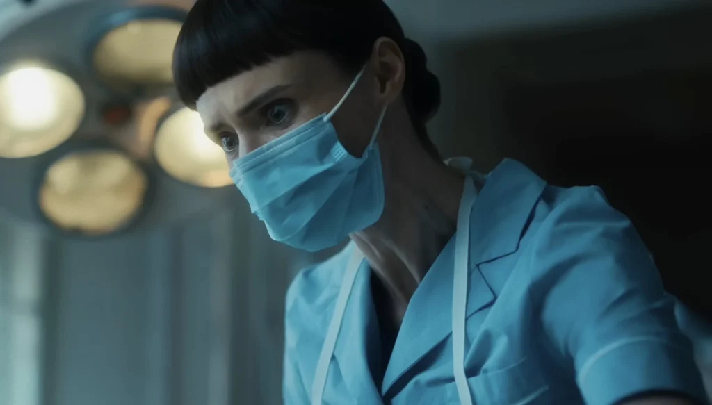 A person wearing a blue medical uniform, apron, and face mask looks intently at something off-camera in a well-lit room.