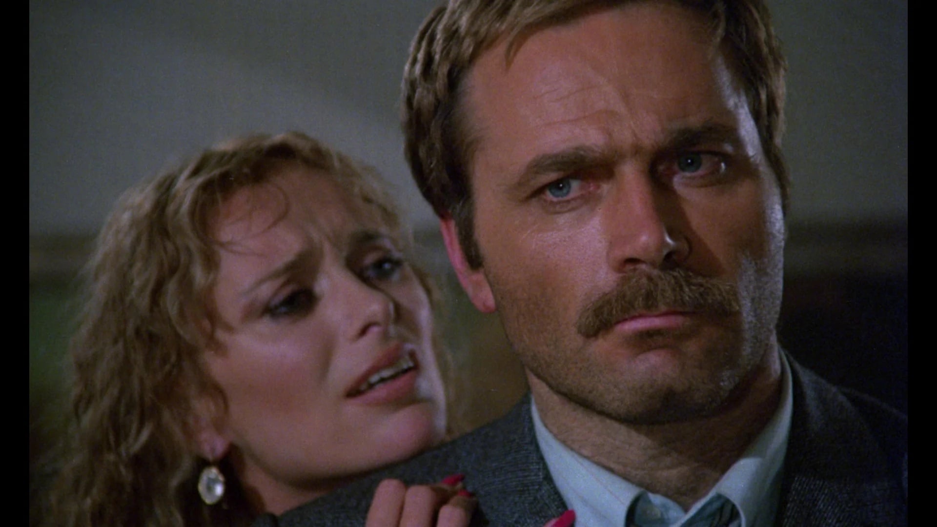 A woman with curly hair and earrings stands close behind a serious-looking man with a mustache, appearing to speak to him while touching his shoulder.