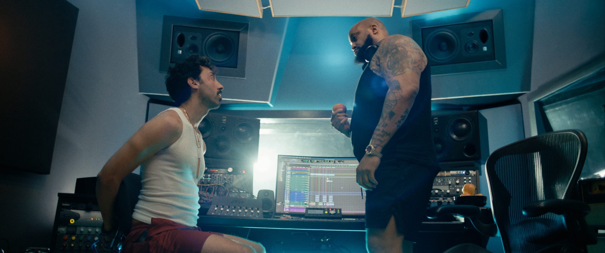 Two men are in a music recording studio; one is seated and the other stands, both engaged in conversation in front of audio equipment and monitors.