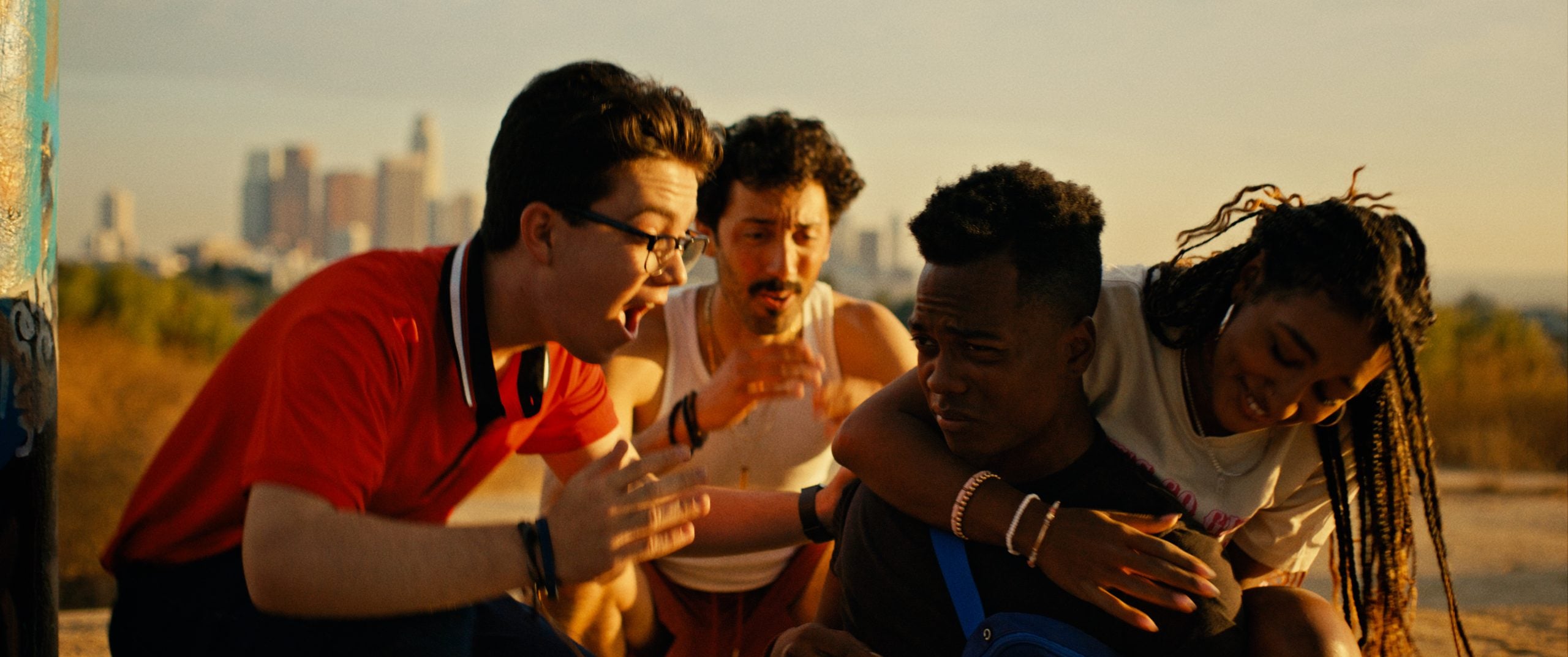 Four young people are outdoors during sunset, with one person crouching and another comforting them while two others gesture animatedly in the background. City skyline is visible in the distance.