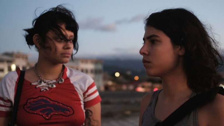Two young people stand outdoors at dusk, looking at each other with serious expressions. One wears a red shirt and necklace, the other wears a striped tank top.