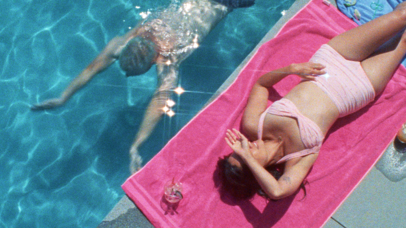 A person swims in a pool while another person lies on a pink towel by the poolside, shielding their eyes from the sun. A drink sits next to the towel.