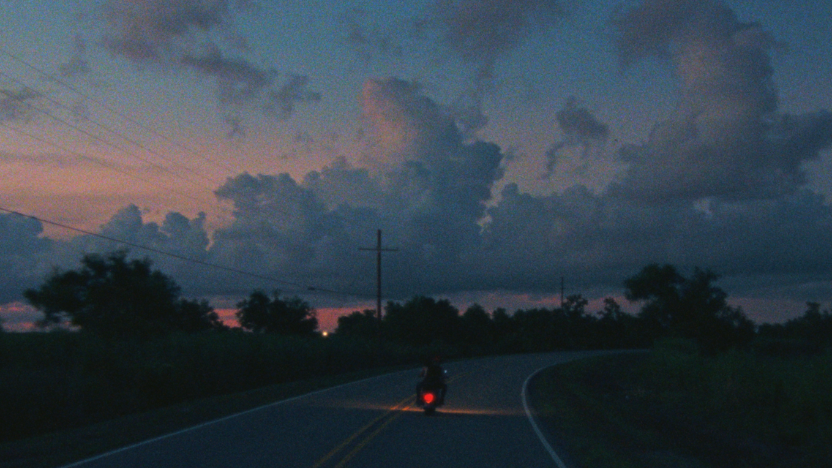 A person rides a motorcycle down a winding road at dusk, with large clouds and a colorful sky in the background.