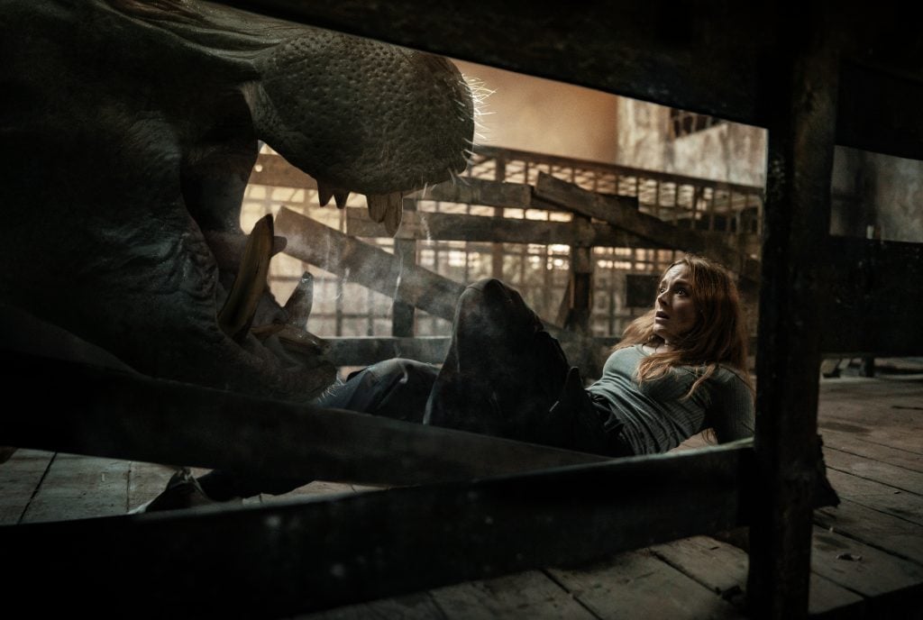 Woman lying under a fallen tree in a dark, abandoned building with a large creature nearby.