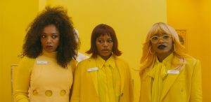 Three women in yellow outfits and name tags stand indoors against a bright yellow background, looking ahead with serious expressions.
