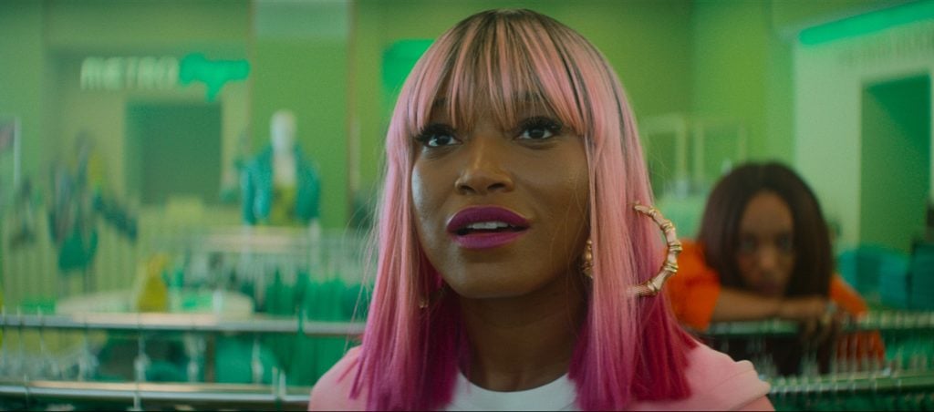 A person with straight pink hair and hoop earrings stands indoors, looking up, with another person leaning on a counter in the blurred background.