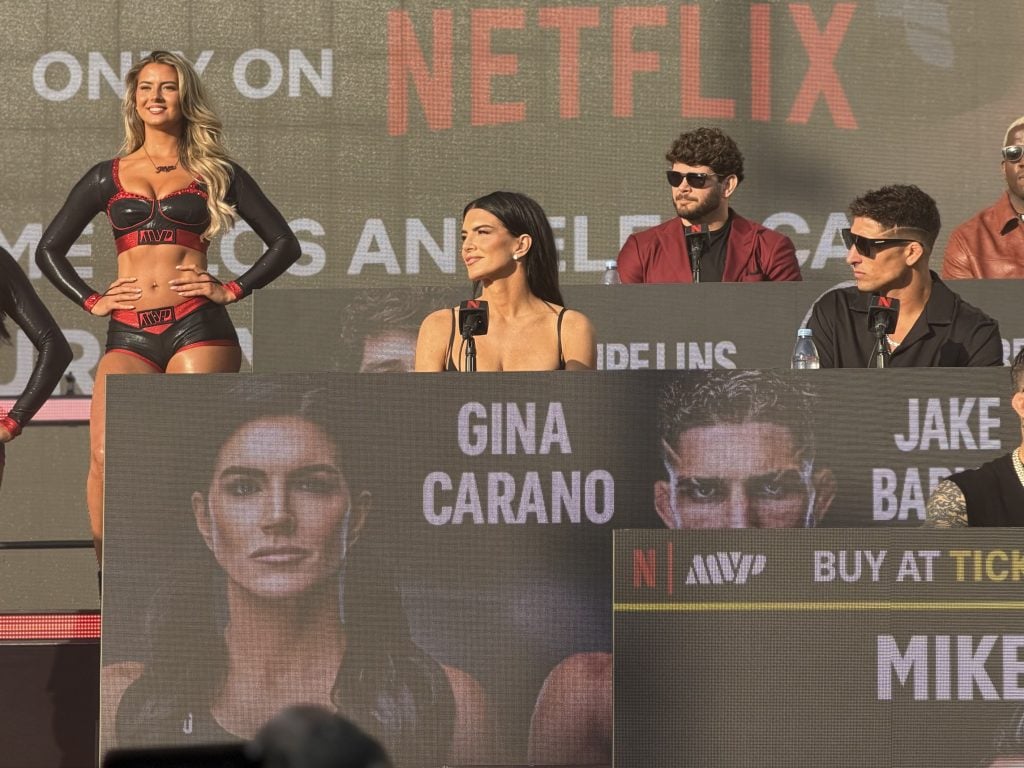 Gina Carano sits at a panel with others, a promotional poster showing her name and face is displayed in front, with Netflix branding visible in the background.