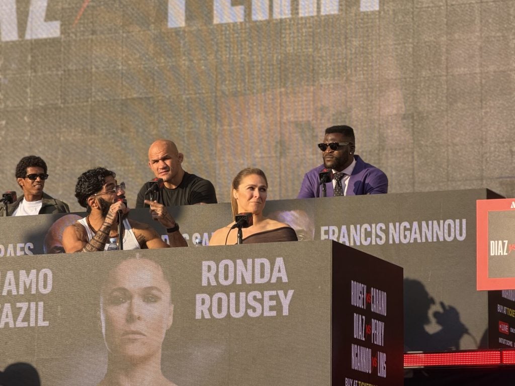 A panel of speakers sits at a press conference table with name cards, including Ronda Rousey and Francis Ngannou, in front of a large screen displaying their names and images.