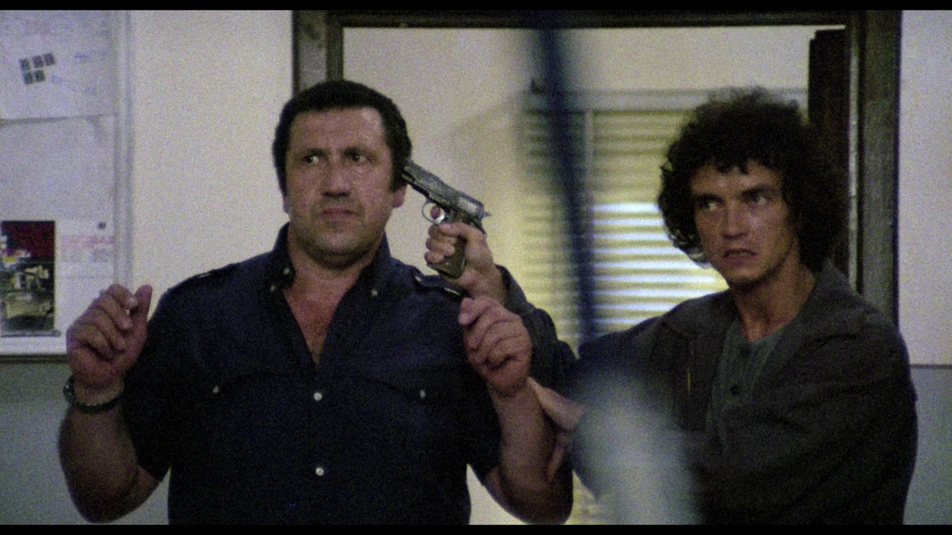A man holds another man hostage at gunpoint inside a room, gripping him by the arm with a revolver pressed to his head.