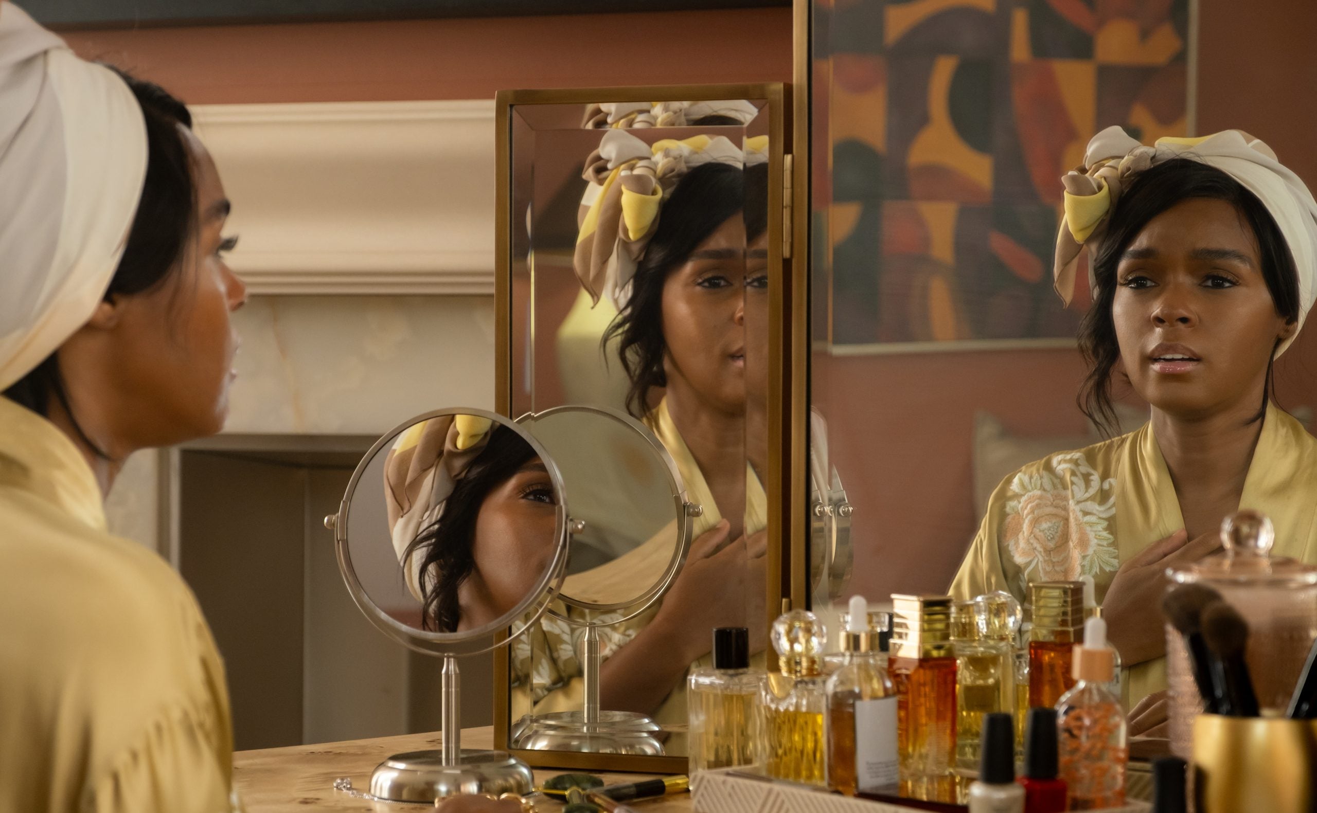 A person wearing a robe and head wrap sits at a vanity table with cosmetics and mirrors, looking at their reflection.