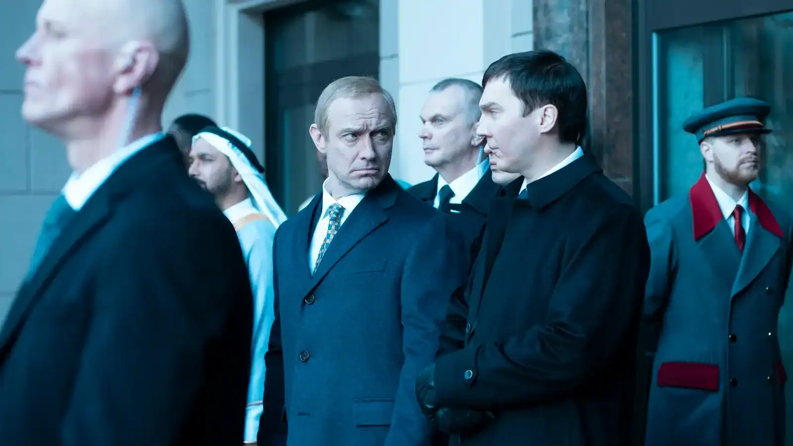 A group of men in formal and traditional attire stand outside a building, engaged in conversation.