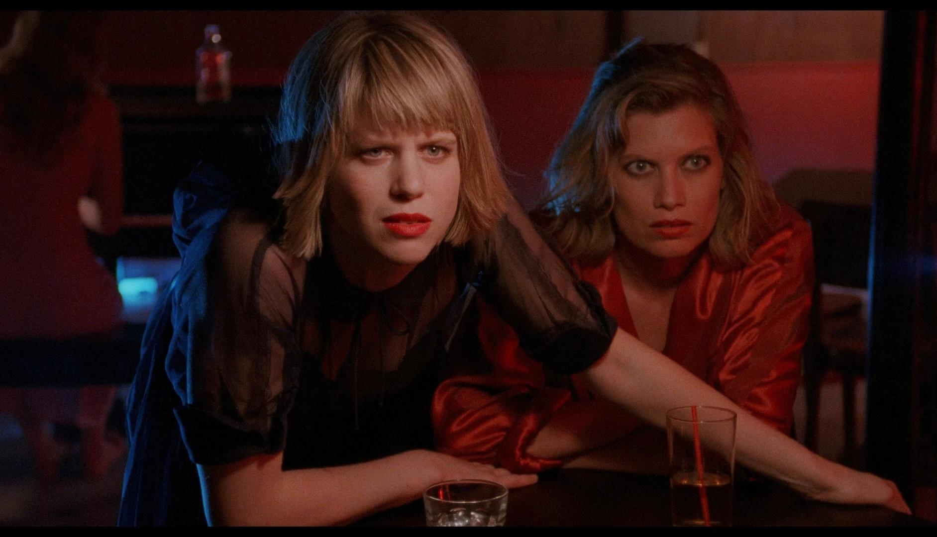 Two women with short blonde hair sit at a dimly lit bar, looking intently ahead. One leans forward with her arm extended, glasses with drinks in front of them.