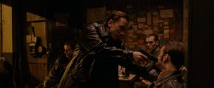 A man in a leather jacket points a gun at another man's head in a dimly lit room, with two other men sitting nearby watching the scene.