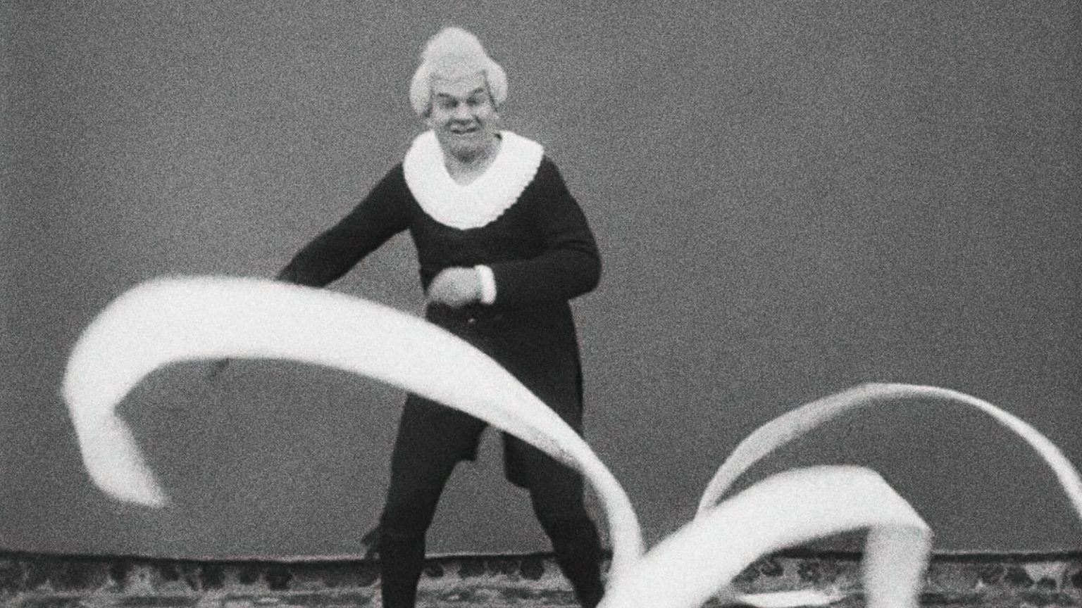 A person in historical costume with a white collar and wig swings long, flowing ribbons in front of a plain background.