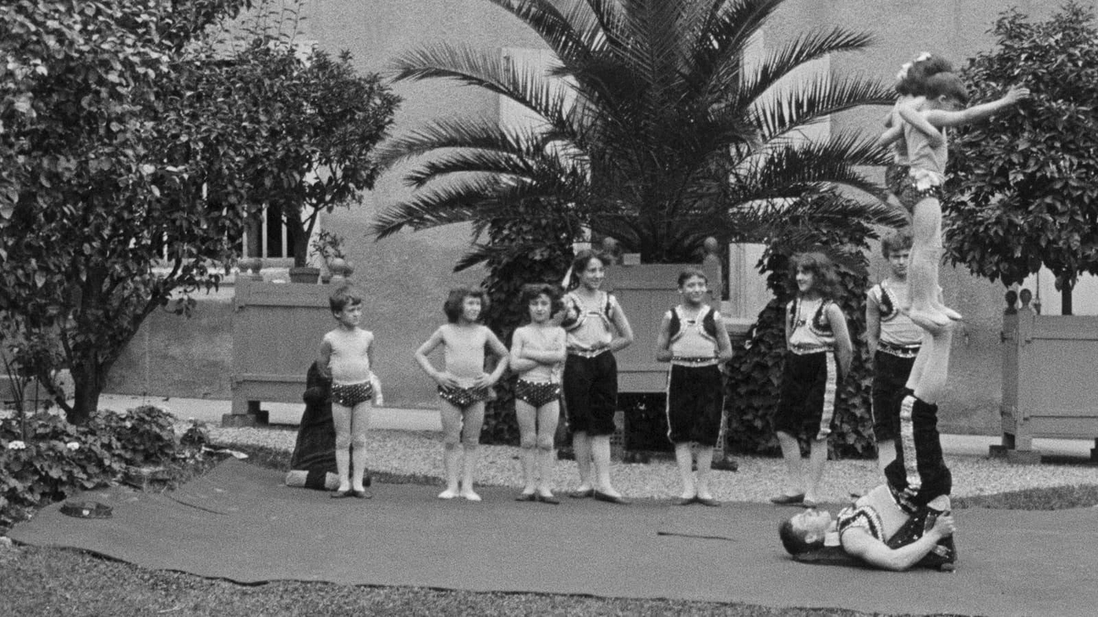 A group of children in costumes stands on a mat outdoors; one child balances another on their feet while lying on their back, with plants and a building in the background.