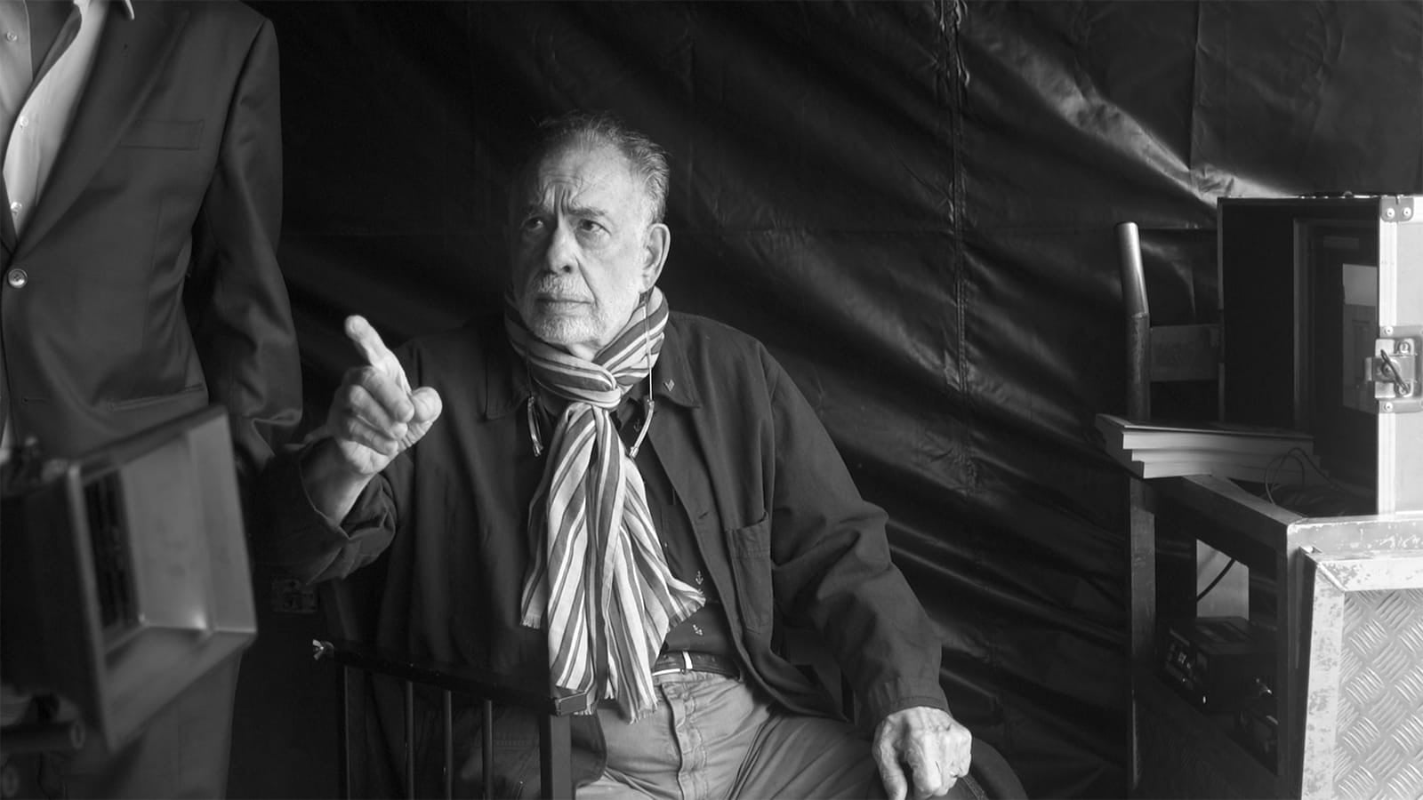 An older man with a striped scarf sits in a chair, gesturing with his finger raised. He appears to be speaking. There is a man standing beside him and technical equipment nearby.