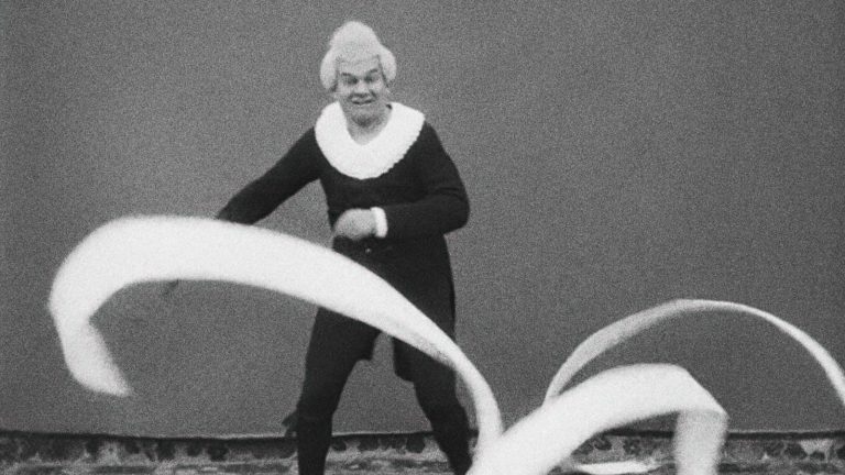 A person in historical costume with a white collar and wig swings long, flowing ribbons in front of a plain background.