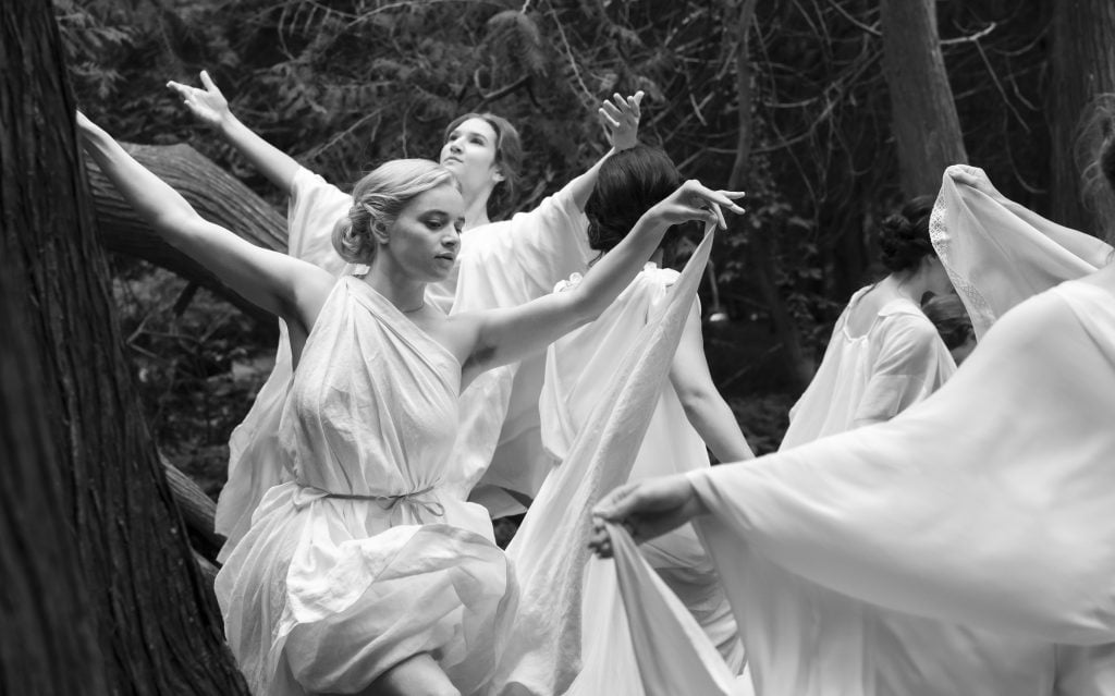A group of women in flowing white dresses dance gracefully among trees in a forest setting.
