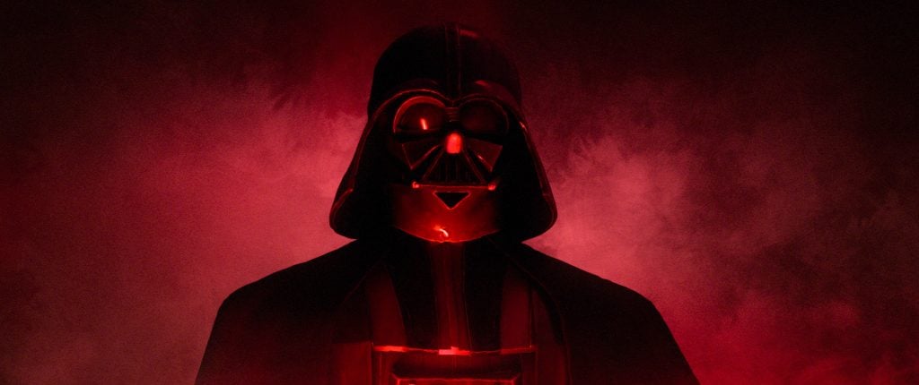 A person dressed as Darth Vader stands against a red, smoky background, illuminated from below.